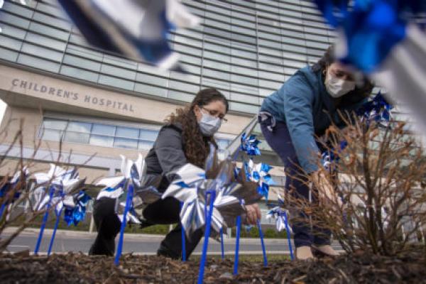 Two women kneel down and place plastic pinwheels into the ground by hand. Several pinwheels are in the foreground, close to the camera. A building with the words “Children’s Hospital” is in the near background.