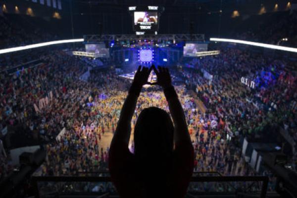 In the foreground a person is silhouetted as they hold up their hands in a 'diamond' formation. THON 2019 at the Bryce Jordan Center is in the background.