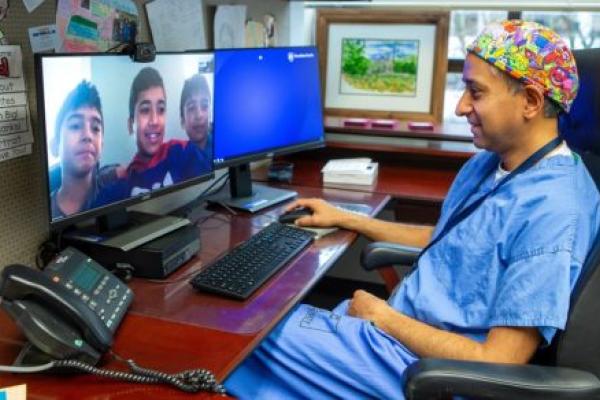 A doctor in scrubs smiles at images of three boys on his computer screen.