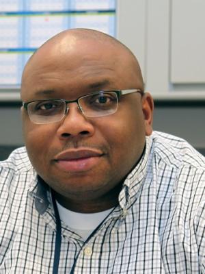 Julius Cabbagestalk, Service Desk team lead, sits at a desk with his hands clasped. He is bald and wears glasses, a checked shirt and a lanyard around his neck.