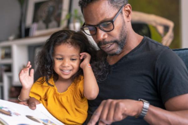 A man holds his young daughter on his lap as they both look down at a picture storybook. A bookshelf, artwork and plants are in the background.