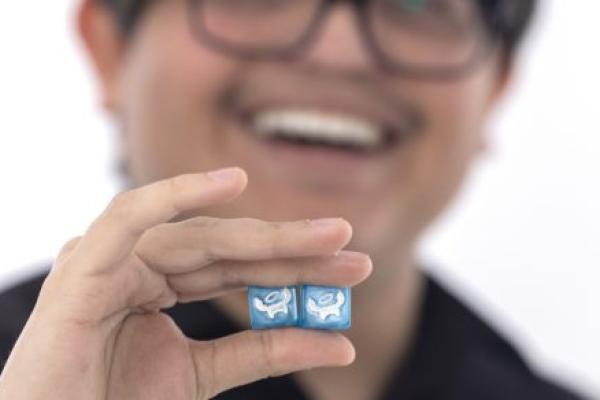 Joseph Romero poses with a pair of specially designed dice that he’s holding near the camera. Joseph is in the near background, slightly out of focus, smiling.