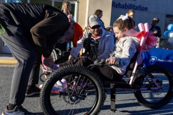 A man leans over to adjust the front of an adaptive bicycle. A young girl sits on the bike smiling, as her dad looks on. Additional people stand in the background.