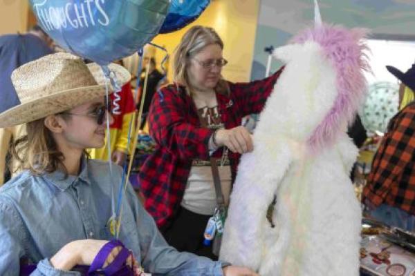 A mother and her teenage son peruse costumes and other items laying on tables. The son, seated in a wheelchair, holds balloons and other items.