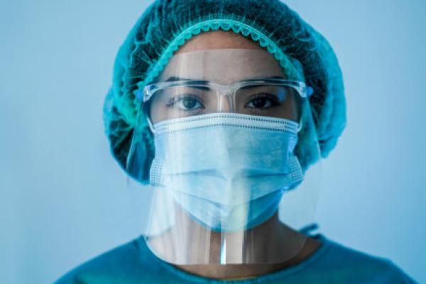 A woman wearing a mask, face shield and other personal protective equipment stands against a wall.