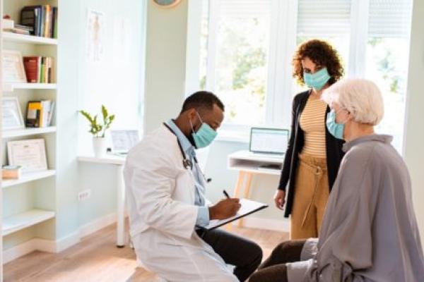 In a doctor’s office, a doctor is seated and writing in a chart while sitting across from a seated, elderly female patient. A younger woman stands by her side. All are wearing face masks.