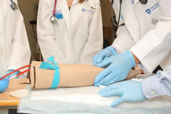 Penn State College of Medicine students work with a simulation arm to learn how to draw blood in a session in the Clinical Simulation Center in July 2016. The simulation arm is pictured in the center of the photo with tubes of red-dyed liquid leading to it. The hands of four students are seen around the simulation arm.