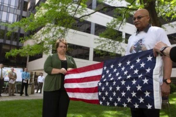 In a courtyard, two people fold an American flag. A man stands at attention in front of the flag. Behind them, a row of people look on from a walkway.